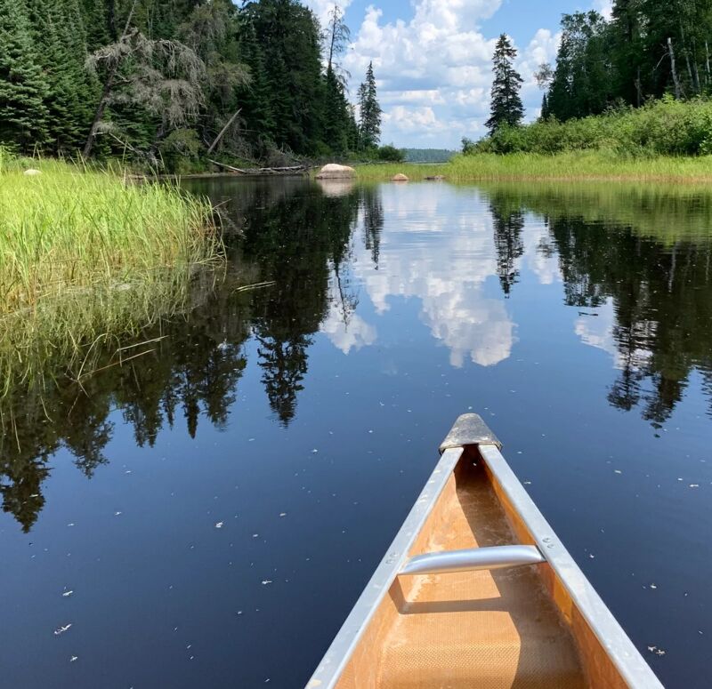 The image shows a canoe on a calm river or lake, surrounded by lush green vegetation and trees. The water reflects the sky and trees, creating a mirror-like effect. The canoe is in the foreground, with the shoreline and trees in the background. The sky is blue with some white clouds, suggesting a peaceful and serene day.